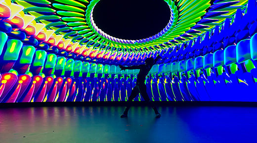 A silhouette of a person in a dance pose on a stage. Behind the person is a screen displaying abstract digital shapes 