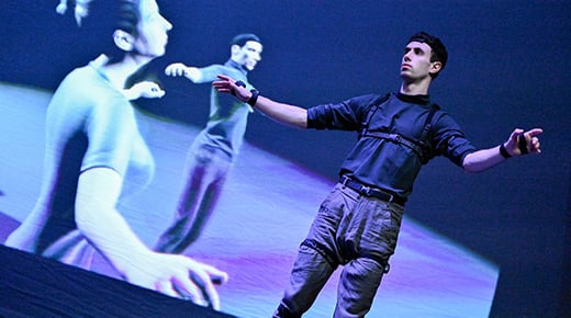 Image credit to Beyond. A dancer stands on stage with their arms outstretched in front of a large screen displaying an avatar of the dancer on stage. A second dancer appears on screen who is not on the stage.