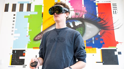 A person wearing virtual reality headset and holding VR controllers stands in front of a wall painted to show a large human eye set over vertical rectangle shapes.