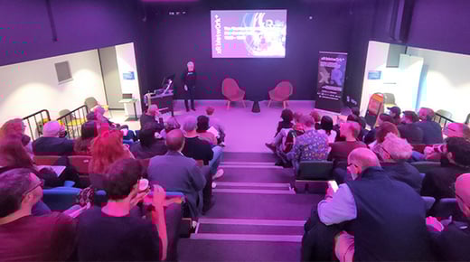 Damian Murphy stands in a lecture theatre at Abertay University in front of an audience sat on tiered seats. A large screen hanging from the wall displays the XR Nework+ logo.