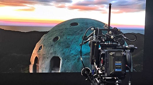 A video camera in the foreground pointing at a large LED volume/screen showing a domed building on a rocky landscape with clouds and sky above
