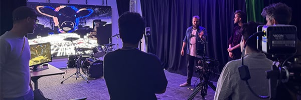 People stand in the XR Stories immersive technology lab facing a large LED wall displaying graphics from digital artist, Danny Bacchus. In front of the large screen are a set of drums and a smaller TV screen on a table. In the foreground a camera points towards the screen.