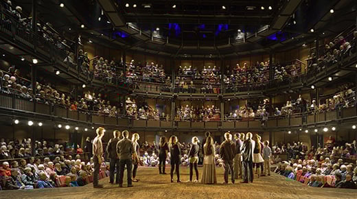 Interior photo of the Royal Shakespeare Theatre auditorium, taken with actors and audience at Builder’s Opening Night. Actors stand on stage at the RSC theatre auditorium facing an audience sat on tiered seats surrounding the stage on three sides. 