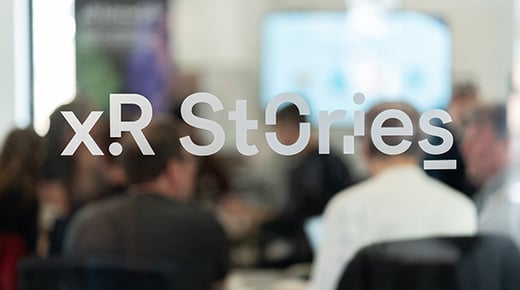 The words XR Stories appear on a glass window. Behind the window an audience sits facing a large TV screen and a presenter.