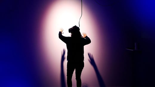 A person wearing a virtual reality headset stands facing a wall with their arms raised as if reaching out to touch something. A light sines behind the person casting a shadow of their hands onto the wall 